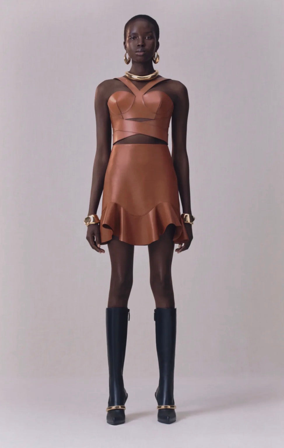 Model wearing a brown leather Alexander McQueen corset dress with black knee-high boots on a plain background