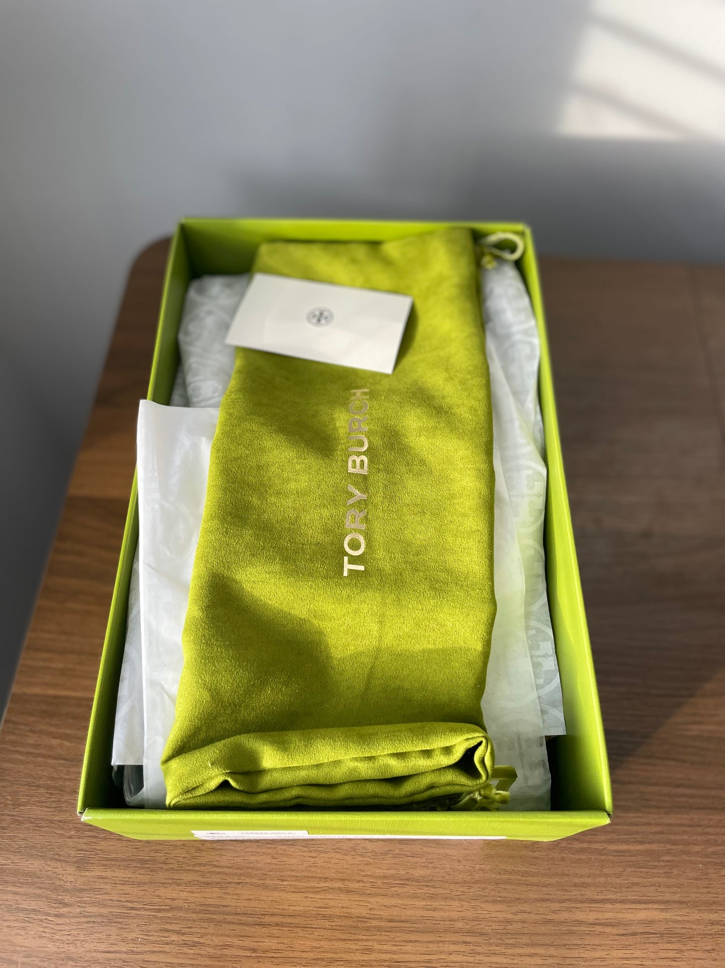 Green folded garment bag with 'Tory Burch' branding inside a green shoe box on a wooden surface.