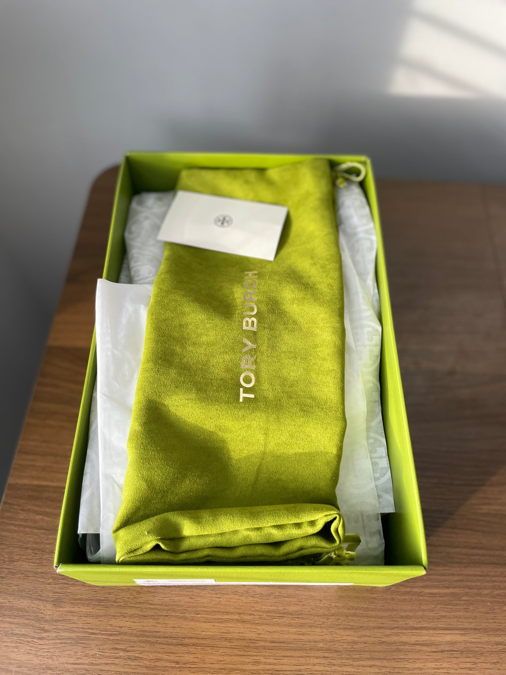 Green folded garment bag with 'Tory Burch' branding inside a green shoe box on a wooden surface.
